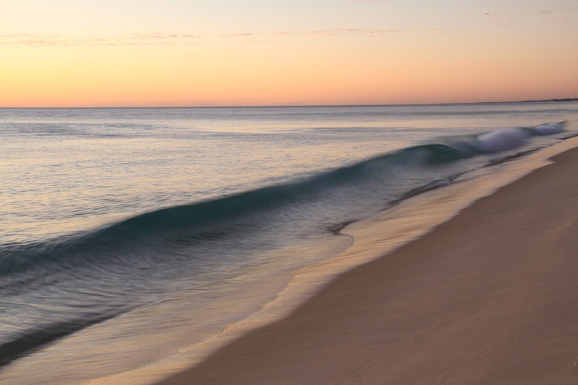 Una ola suave llegando a una playa vacía bajo un cielo melocotón